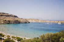 Lindos.  Shallow bay with tourists swimming and moored boats  with line of sun umbrellas along curve of beach in high summer season.Greek IslandsAegeanRodicoast coastalseaswimbathingblue skyc...