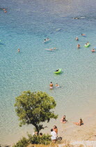 Lindos.  Shallow bay with tourists swimming and sitting on beach in high summer season.  Single tree in foreground.Greek IslandsAegeanRodicoast coastalseaswimbathingblue skyclearcloudlessre...