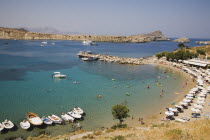 Lindos.  Shallow bay with tourists swimming and moored boats with lines of sun umbrellas following curve of beach in high summer season.Greek IslandsAegeanRodicoast coastalseaswimbathingblue s...