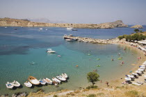 Lindos.  Shallow bay with tourists swimming and moored boats with lines of sun umbrellas following curve of beach in high summer season.Greek IslandsAegeanRodicoast coastalseaswimbathingblue s...