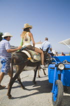 Lindos.  Classic Greek Island scene with tourists on donkey being led from the Akropolis of Lindos down to beach past bright blue delivery motorbike on paved stone path with whitewashed church part se...