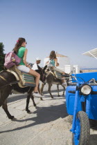 Lindos.  Classic Greek Island scene with tourists on donkey being led from the Akropolis of Lindos down to beach past bright blue delivery motorbike on paved stone path with whitewashed church part se...