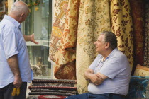 Two men in conversation outside shop display of rugs and carpets.  One seated  one standing  facing each other.AegeanGreek IslandsRhodiSummertalk talkingcoast coastalresortholidaypackagetrip...