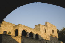 Exterior walls and colonnade of stone building framed by silhouetted archway in late afternoon sunshine.AegeanGreek IslandsRhodiSummercoast coastalresortholidaypackagetripDestination Destina...