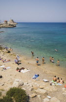 Tourist sunbathers and swimmers on rocky beach beside shallow stretch of aquamarine sea with view towards medieval stone windmills on harbour wall beyond.AegeanGreek IslandsRhodiseaSummercoast c...