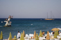 Rhodes Town.  Diving platform in sea with boys queueing to jump and swimming around base with lines of striped parasols and sunbeds along beach in foreground and day trip boat in distance behind.Aege...