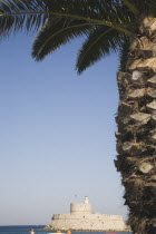 Rhodes Town.  Circular fort at entrance to Mandraki harbour from beneath palm tree on town beach part seen in foreground.AegeanGreek IslandsRhodiSummerseacoast coastalmedieval fortified fortif...