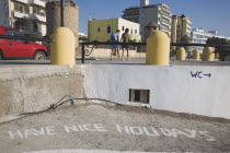 Rhodes Town.  Slogan Have Nice Holidays painted on entrance steps to beach beside wall with painted direction to WC.  Road and town buildings beyond.AegeanGreek IslandsRhodiSummerseacoast coasta...