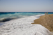 Rhodes Town.  Young couple in swimwear walking along Rhodes Town beach beside white  breaking surf and clear  shallow  aquamarine water beyond.AegeanGreek IslandsRhodiSummerseacoast coastalreso...