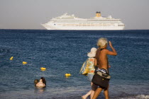 Rhodes Town.  Middle aged couple walking along town beach with cruise ship on horizon behind and tourists in water. AegeanGreek IslandsRhodiSummerseacoast coastalresortholidaypackagetripDes...