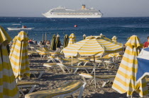 Rhodes Town.  Yellow and white striped parasols and sun loungers on Rhodes Town beach with cruise ship on the horizon behind. AegeanGreek IslandsRhodiSummerseacoast coastalresortholidaypackag...