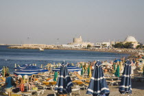 Rhodes Town.  Striped parasols and sun loungers on busy town beach with fort at entrance to Mandraki harbour behind.AegeanGreek IslandsRhodimedievalfortified fortificationsSummerseacoast coast...