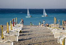 Rhodes Town.  Lines of rental sunbeds and parasols packed and folded on Rhodes Town beach at the end of the day with family sitting on sand and small craft sailing boats as students learn to sail on e...
