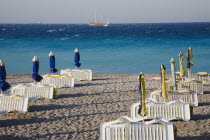Rhodes Town.  Lines of rental sunbeds and parasols packed and folded on Rhodes Town beach at the end of the day with expanse of clear  aquamarine water behind and distant cruise boat returning to harb...