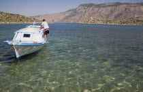 Boat moored in clear shallow water off Ag. Panormitis monastery in the bay on the south of the island with male figure walking along side.AegeanGreek IslandsSimiharbour Summerseacoast coastalr...