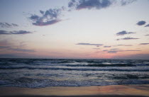 Kusadasi.  Shoreline at sunset looking west to nearby Greek island of Samos with pale pink sky reflected in wet surface of sand.waveshallowreflectioncloudeveninglighttwilightdusk Blue Destinat...