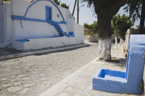 Pythagorio.  Cobbled street with blue and white painted church exterior and blue painted roadside shrine on right.  Tree with lower half of trunk also painted white.AegeanGreek IslandsPithagorion P...