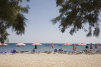 Pythagorio.  Line of red and blue beach parasols and sun loungers with early season tourists and flat calm sea beyond.  Part framed by tree branches in foreground.North Eastern AegeanGreek IslandsP...