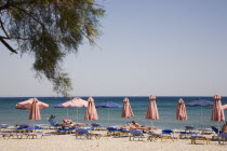Pythagorio.  Line of red and blue beach parasols and sun loungers with early season tourists and flat calm sea beyond.  Overhanging tree branches in foreground.North Eastern AegeanGreek IslandsPith...