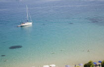 Pythagorio.  Yacht moored in the bay with tourists swimming in clear  aquamarine water in early Summer season and line of parasols along beach partly seen in foreground.North Eastern AegeanGreek Isl...