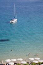 Pythagorio.  Yacht moored in the bay with tourists swimming in clear  aquamarine water in early Summer season and line of white parasols along beach in foreground.North Eastern AegeanGreek IslandsP...