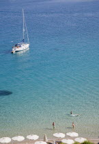 Pythagorio.  Yacht moored in the bay with tourists swimming in clear  aquamarine water in early Summer season and line of white parasols along beach in foreground.North Eastern AegeanGreek IslandsP...