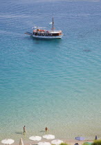 Pythagorio.  Tourists swimming in clear water in early Summer season with day trip boat returning late afternoon from island tour and line of parasols on beach part seen in the foreground.North Easte...