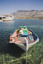 Greek painted fishing boat moored in clear  shallow water off St Michael Panormitis monastery with ferry boat moored at jetty behind and headland of southern Symi beyond. AegeanGreek IslandsSimico...