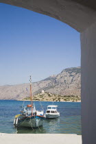 Pleasure boat and fishing boat moored in shallow water and coastline behind framed by archway in St Michael Panormitis monastery.AegeanGreek IslandsSimicoast coastalseaSummerpackageholidayres...