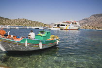 Green and white painted fishing boat moored in clear  shallow water off St Michael Panormitis monastery with ferry moored on jetty behind.AegeanGreek IslandsSimicoast coastalseaSummerpackageho...