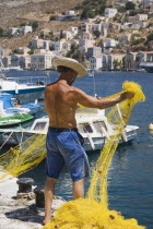 Fisherman from Symi in shorts and straw sun hat preparing nets on jetty with whitewashed houses of Yialos harbourfront behind.AegeanGreek IslandsSimicoast coastalseaSummerpackageholidayresort...