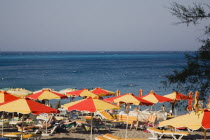 Yialos.  Red and yellow striped sun umbrellas and sun loungers on beach with expanse of calm  blue sea beyond.AegeanGreek IslandsSimicoast coastalseaSummerpackageholidayresortvacationtripd...