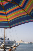 Yialos.  Colourful  striped parasol part seen in foreground of view across water with moored pleasure boats towards stone wall and tower.AegeanGreek IslandsSimicoast coastalseaSummerpackagehol...