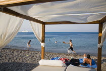 Yialos.  People playing bat and ball game  swimming and sunbathing on peeble beach with view across water towards distant cruise ship framed by canopy of bed draped with white material.AegeanGreek I...