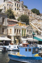 Yialos.  Blue painted fishing boat moored in harbour with line of pleasure boats behind overlooked by pastel coloured facades of waterfront houses.AegeanGreek IslandsSimicoast coastalseaSummerp...