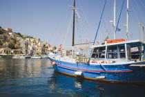 Yialos.  Blue painted fishing boat moored in flat calm water with waterfront buildings on rocky hillside behind.AegeanGreek IslandsSimicoast coastalseaSummerpackageholidayresortvacationtrip...