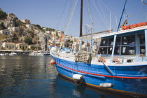 Yialos.  Blue painted fishing boat with red trim moored in flat calm water with waterfront buildings on rocky hillside behind.AegeanGreek IslandsSimicoast coastalseaSummerpackageholidayresort...