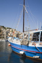 Yialos.  Blue painted fishing boat with red trim moored in flat calm water with waterfront buildings on rocky hillside behind.AegeanGreek IslandsSimicoast coastalseaSummerpackageholidayresort...