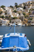 Yialos.  Small  covered  blue painted boat moored in foreground with waterfront buildings extending over steep  rocky hillside beyond.AegeanGreek IslandsSimicoast coastalseaSummerpackageholida...