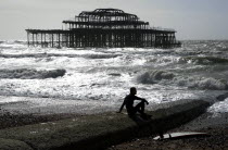 England, East Sussex, Brighton, Seafront, man sat with surf board on goyne in front of the burnt out shell of the former West Pier.