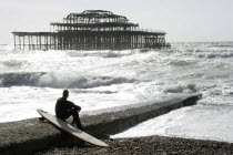England, East Sussex, Brighton, man sat with surf board on goyne in front of the burnt out shell of the former West Pier.