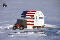 USA, New Hampshire, Harrisville, ice fishermen on Silver Lake. Hut with stars & stripes flag and snow chains on motor trike. Live Free or Die message painted on entrance to Cabin.