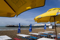 England, East Sussex, Brighton, The Pier at low tide with shingle pebble beach in foreground seen from beneath sunshade umbrellas and empty tables on the seafront.