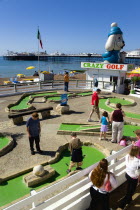 England, East Sussex, Brighton, Adults and children playing miniature golf on a Crazy Golf course on the promenade with Brighton Pier and the pebble shingle beach beyond.