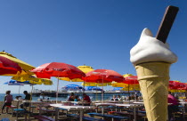 England, East Sussex, Brighton, The Pier with people under sunshade umbrellas by tables on the seafront with a fibreglass ice cream cone in the foreground