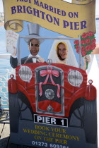 England, East Sussex, Brighton, Man and woman with their faces in amusement cut-out of wedding car that says Just Married On Brighton Pier which also serves as a bookings advertisement for marriage ce...