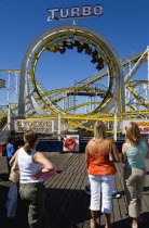 England, East Sussex, Brighton, Women watching the Turbo rollercoaster amusement ride on Brighton Pier with the cars full of people upside down at the top of a loop.