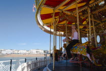 England, East Sussex, Brighton, The Pier with a happy laughing young girl on a traditional Victorian galloping horses fairground carousel roundabout ride with the beach and seafront buildings in the d...