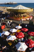 England, East Sussex, Brighton, People sitting under sun shade umbrellas at tables on the promenade outside bars and restaurants with a traditional Victorian galloping horses carousel fairground round...