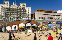 England, East Sussex, Brighton, Young people playing beach volleyball on sand on the seafront with the De Vere Grand Hotel and The Brighton Centre beyond.